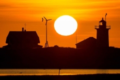 A lighthouse on the beach, a wind turbine and the setting sun.