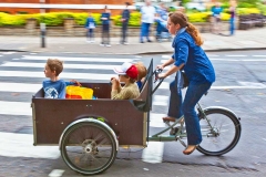 A woman riding a bicycle with a cart attached carrying children.