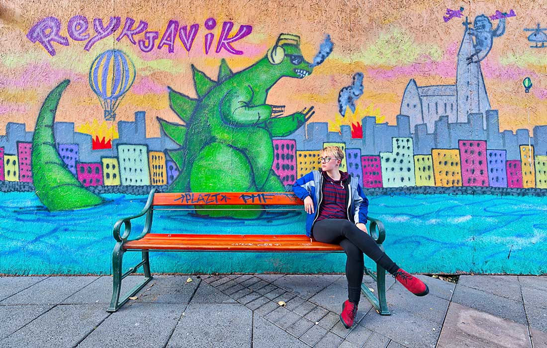 Young woman sits on bench in front of painting with Dinosaur in Reykjavik, Iceland.