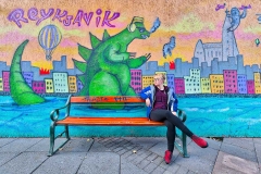 Young woman sits on bench in front of painting with Dinosaur in Reykjavik, Iceland.